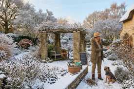 Home gardener pruning plants in a snow-covered winter garden with pergola and cottage during seasonal garden maintenance.