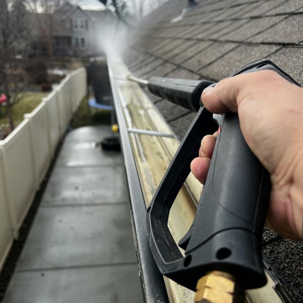 Person using a high-pressure washer to clean residential roof gutters, removing dirt and debris for effective gutter maintenance.