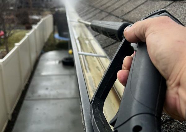 Person using a high-pressure washer to clean residential roof gutters, removing dirt and debris for effective gutter maintenance.
